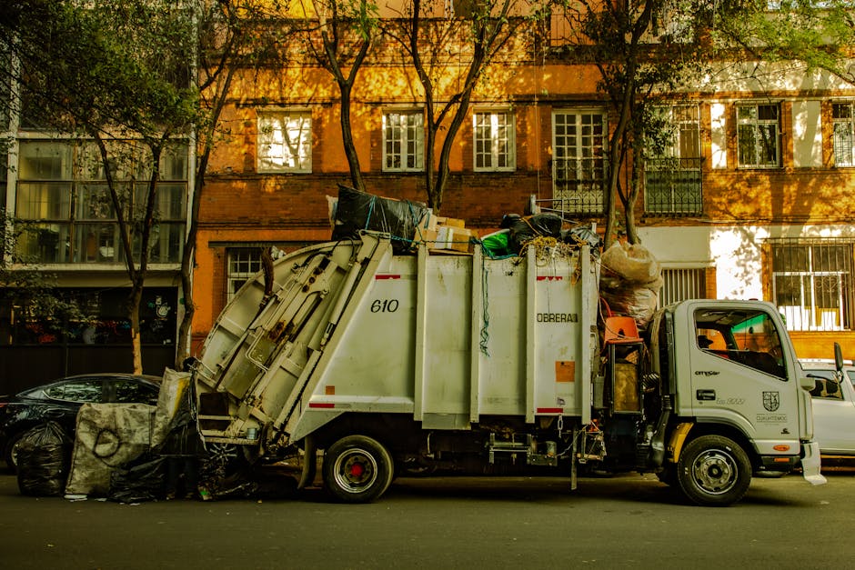 An outdoor scene showing a large collection of household waste and recycling bins overflowing with rubbish, including black, black with white labels, and red bins, as well as open cardboard boxes and