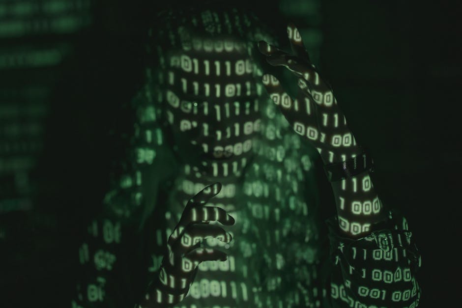 A person with their face and hands illuminated by green digital binary code projected onto their skin and clothing, set against a dark background. The individual appears to be in a contemplative pose,