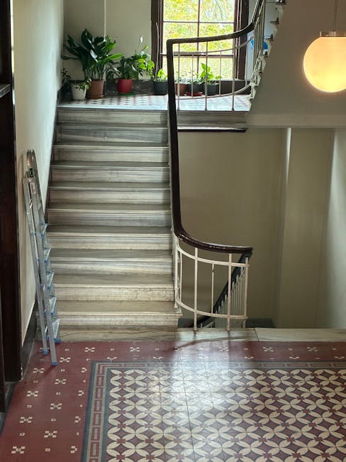 Inside a residential property, a wooden staircase with a curved metal handrail leads from an upper floor down to a tiled landing area decorated with a geometric patterned border. Natural light filters